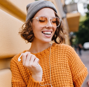 A girl in a sweater and hat is listening to music on her smartphone, smiling.